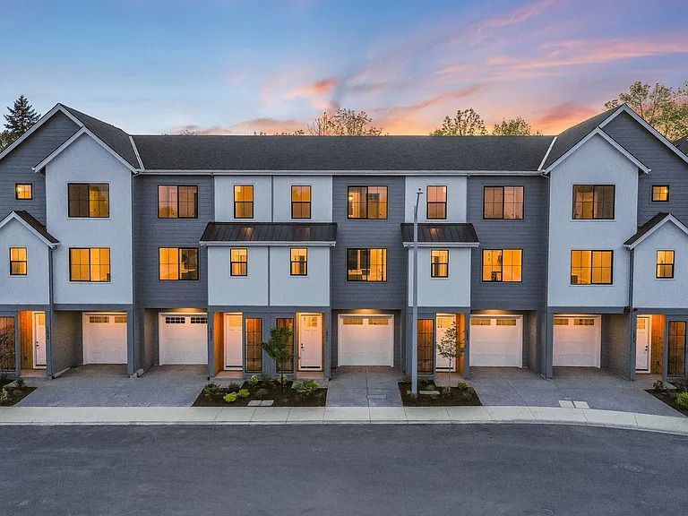 Row of modern townhouses with gray and white exteriors, lit windows, and attached garages at dusk.