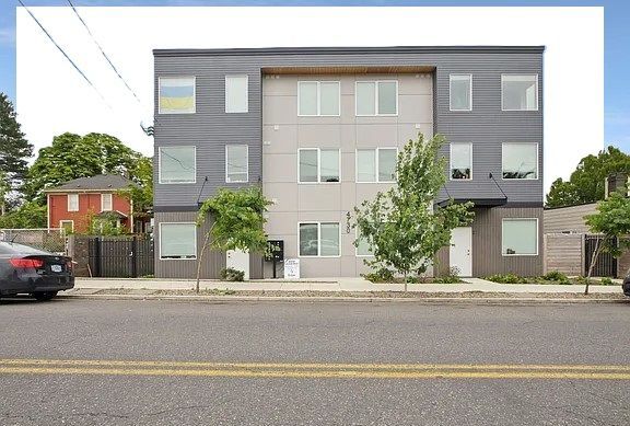 Modern three-story apartment building with gray and light gray panels and white-framed windows, street view.