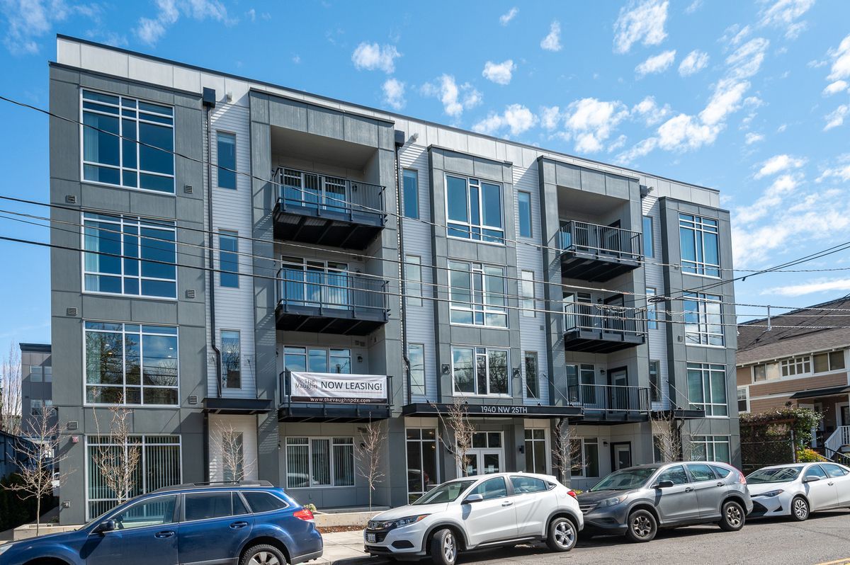 Modern gray apartment building with balconies, cars parked in front, blue sky.