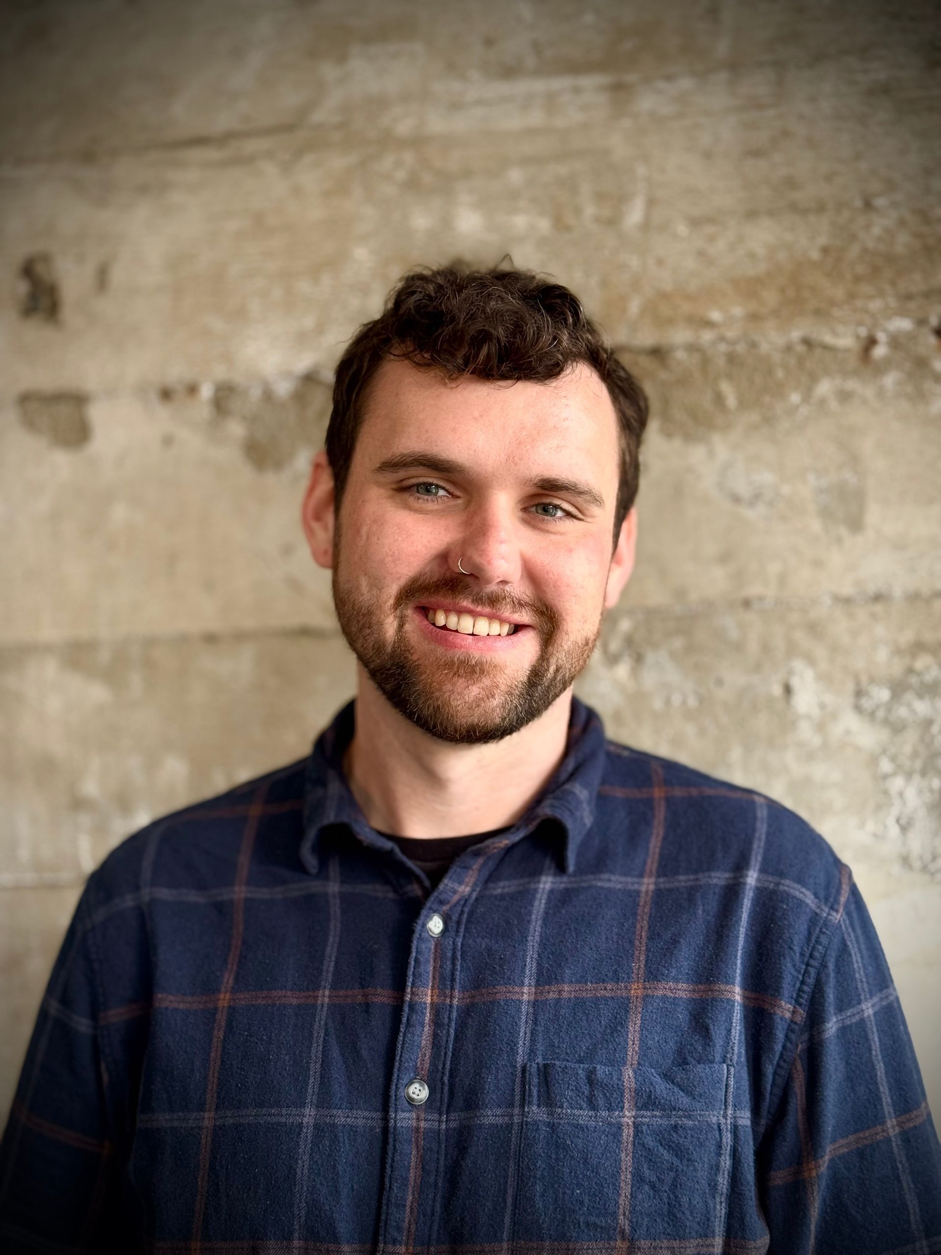Man with short brown hair and beard smiles, wearing a blue plaid shirt, in front of a textured gray wall.