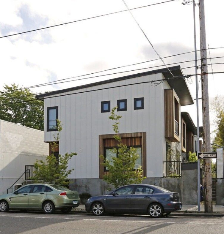 Modern white and wood-trimmed house with cars parked in front. Black square windows, small trees.