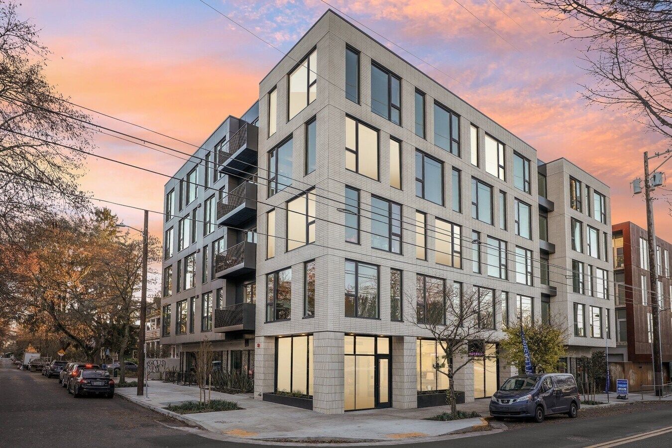 Modern multi-story building at a street corner with large windows and a sunset sky.