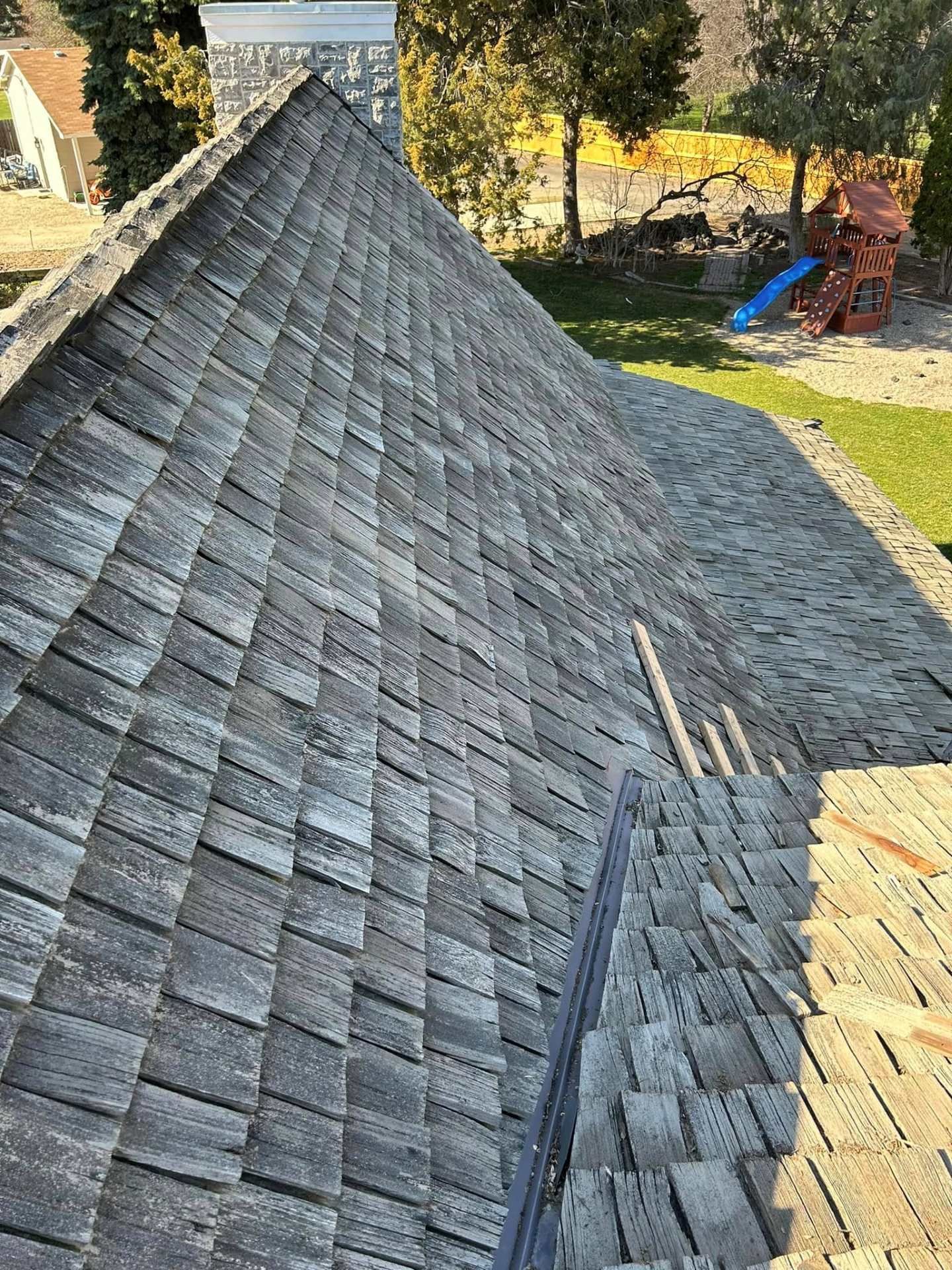 Roof shingles over a steeply pitched roof in a suburban yard, with trees and a playset in the background