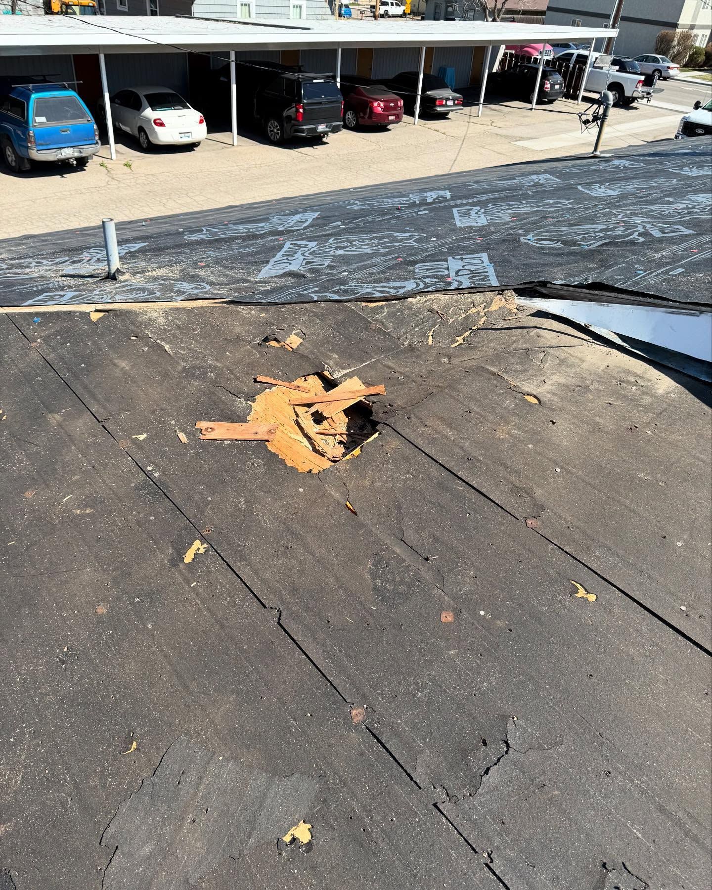 Damaged flat rooftop with a torn hole exposing orange underlayment near parked cars below.
