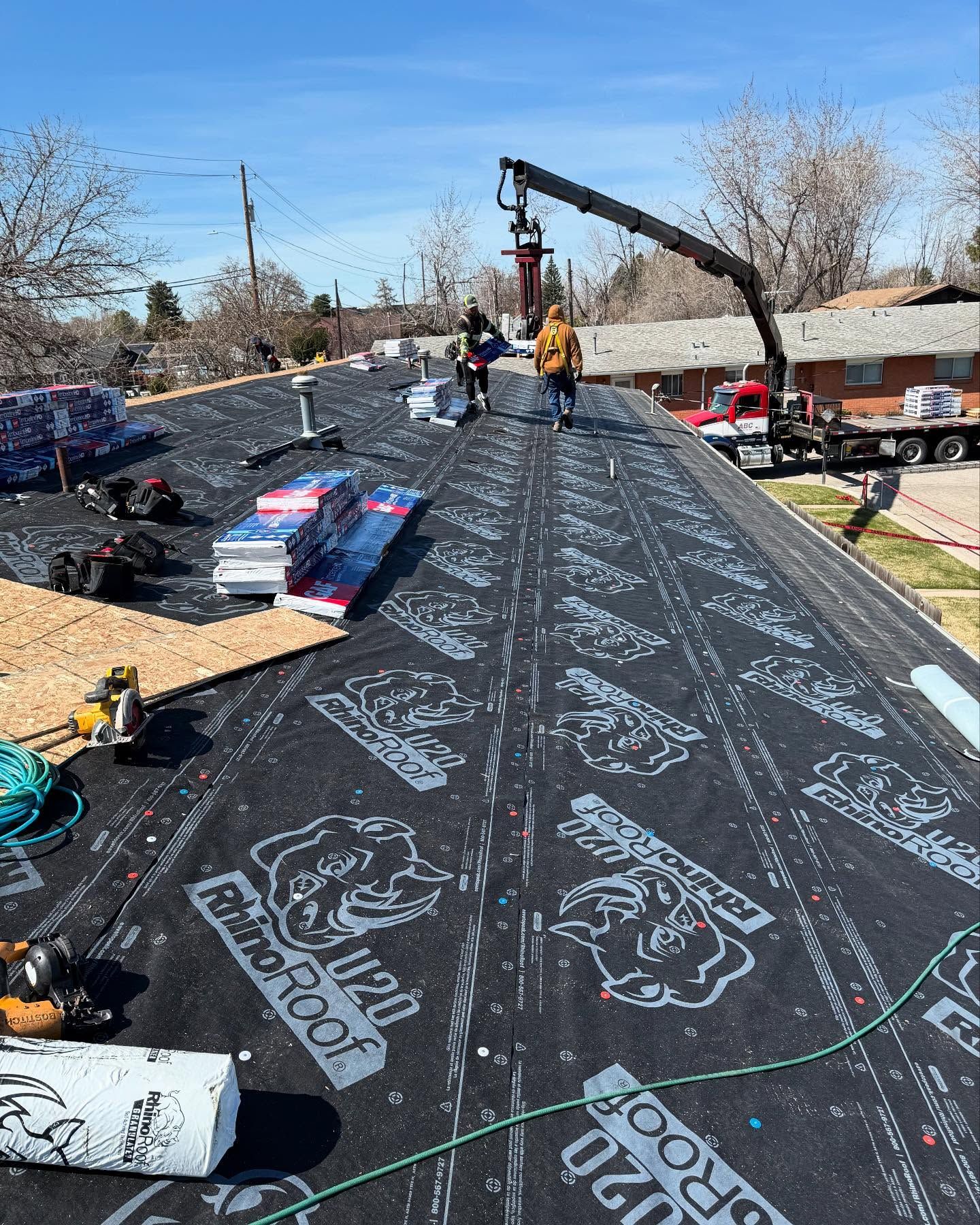 Workers installing black roofing underlayment with a crane on a sloped roof under a blue sky