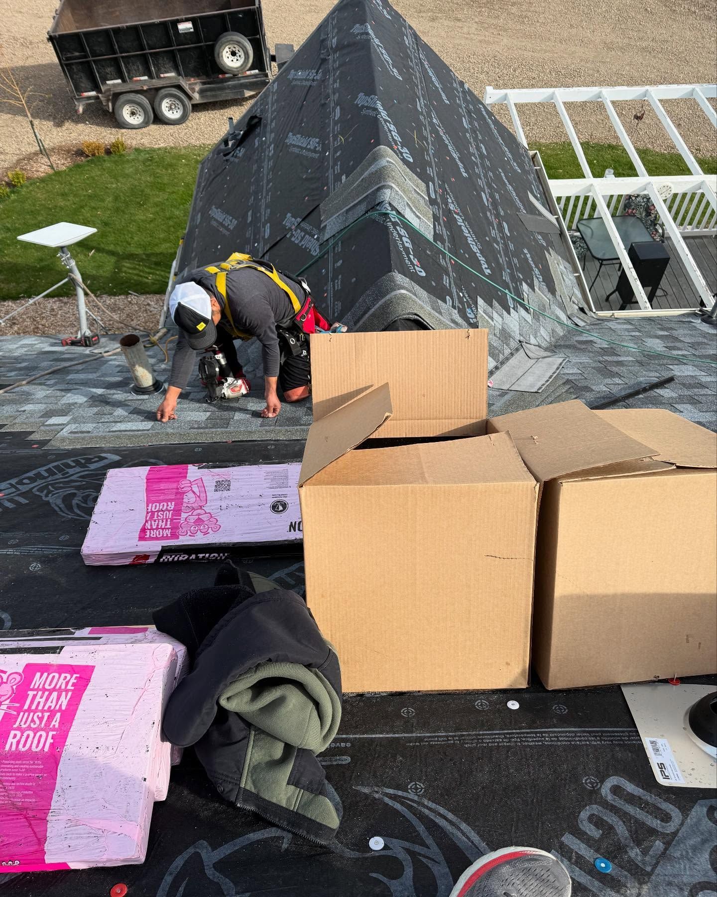 Workers installing roofing on a steep black roof beside stacked cardboard boxes and pink insulation boards