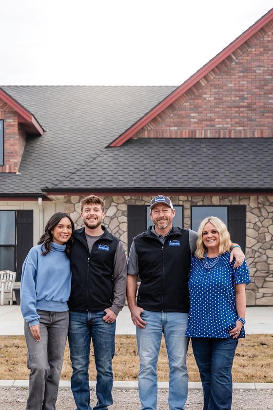 Four people stand smiling in front of a brick house with a shingled roof.