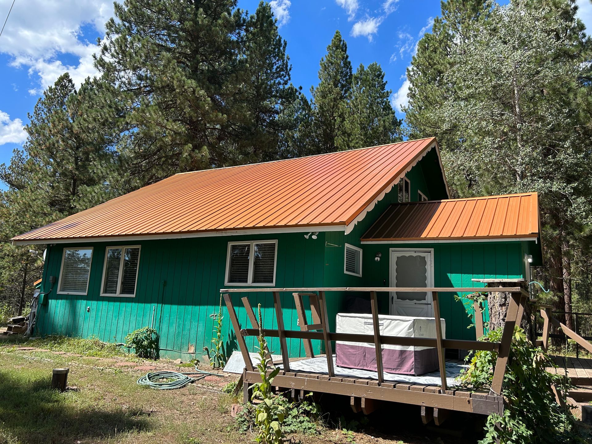 Green cabin with orange roof in a wooded yard, with a small deck under a blue sky.