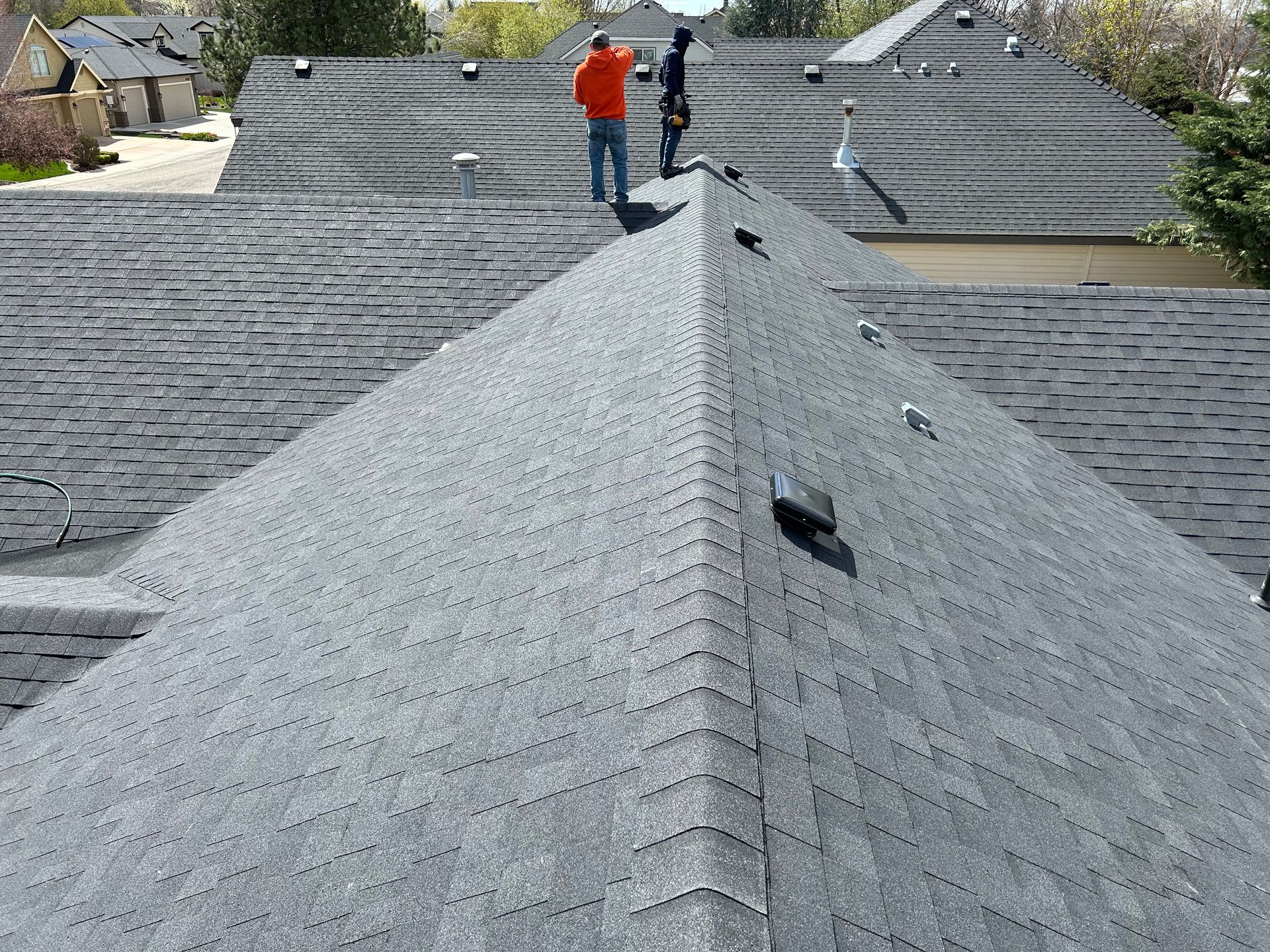 Workers on a gray shingled roof, inspecting or repairing it on a sunny day.