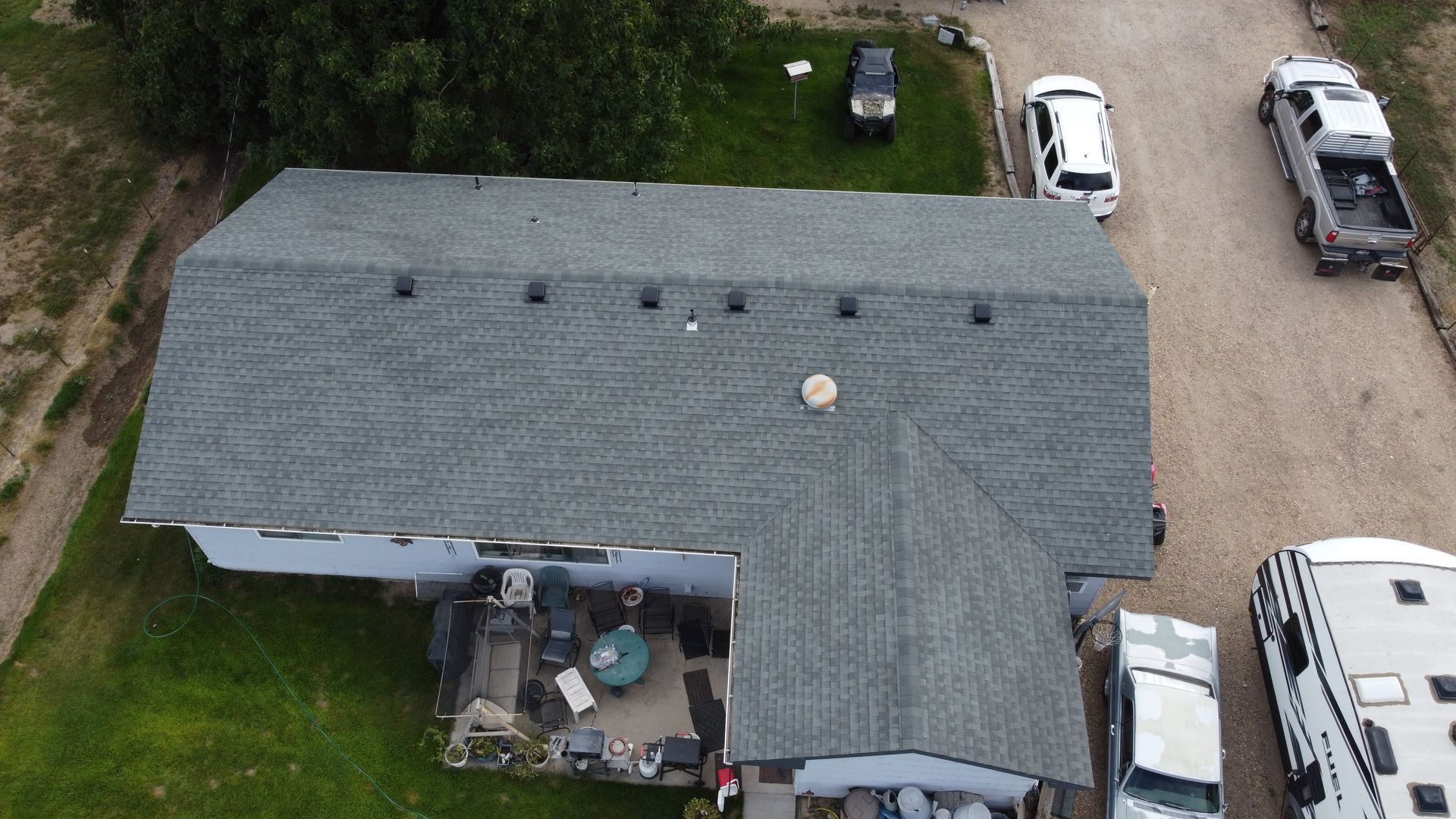 Aerial view of a gray-roofed house with a patio, parked cars, and gravel driveway surrounded by trees.