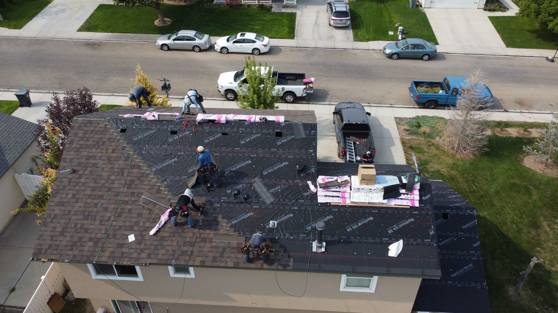 Workers repairing a dark shingle roof on a house, with materials and vehicles on the street below.