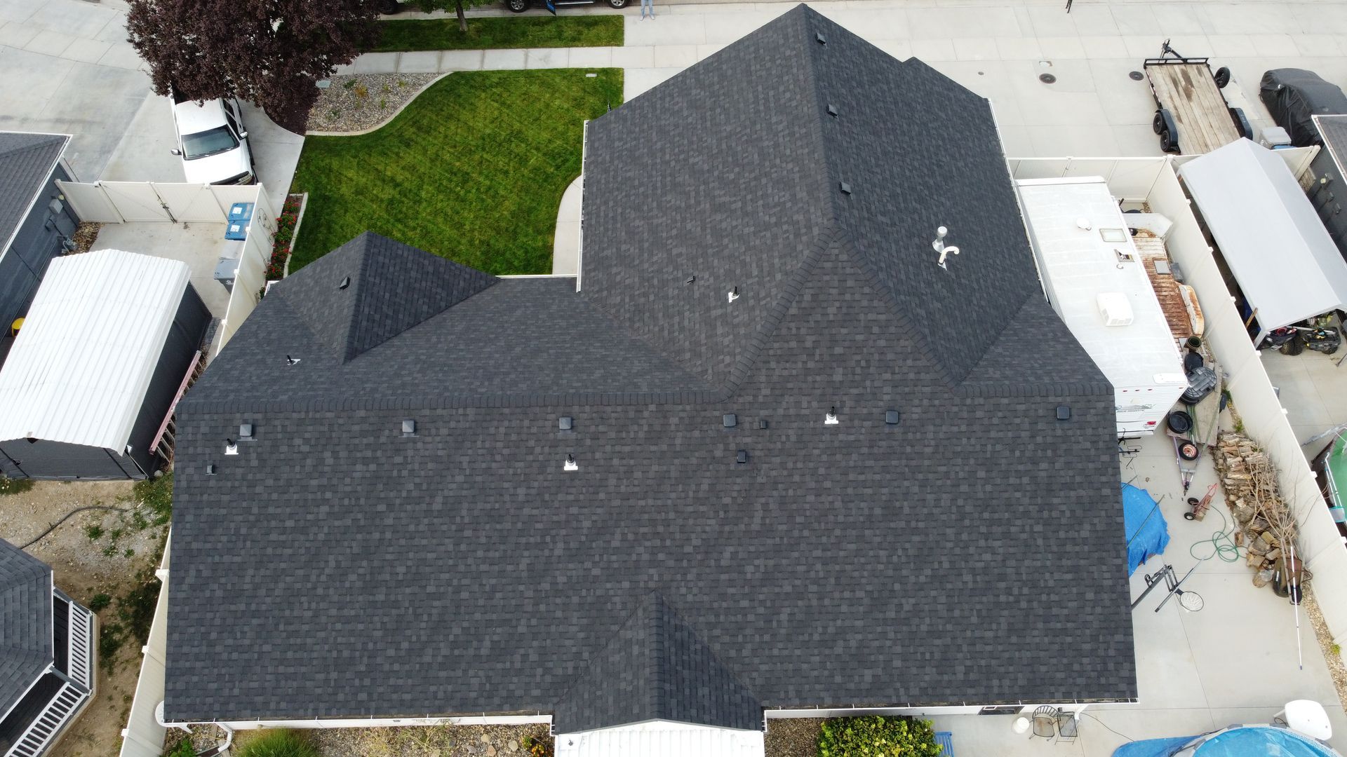 Aerial view of a dark gray shingled house roof with a small green lawn and neighboring structures around it.