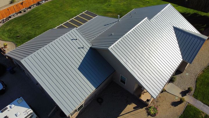 Aerial view of a house with a gray metal roof and solar panels beside a paved driveway.