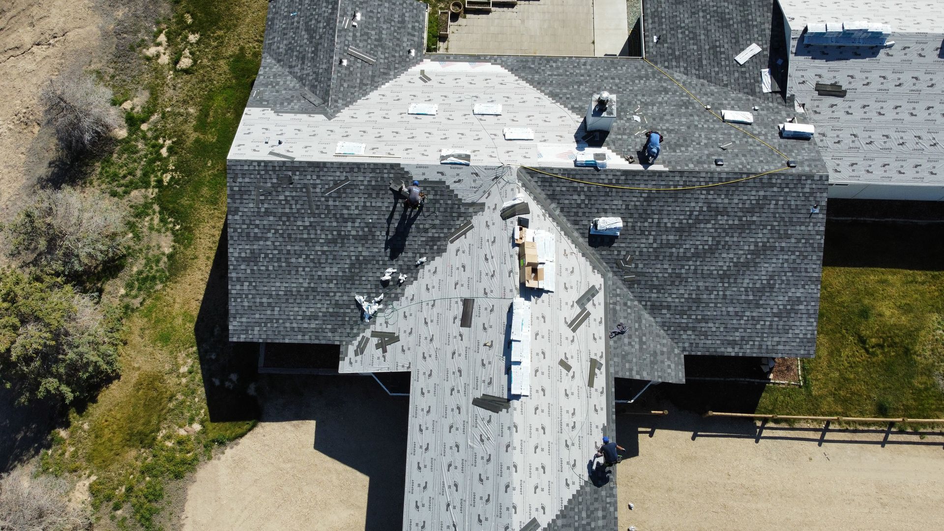 Aerial view of a house roof under construction with workers, dark shingles, and exposed underlayment.