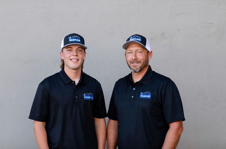 Two men in black polos and blue caps standing against a plain gray wall.