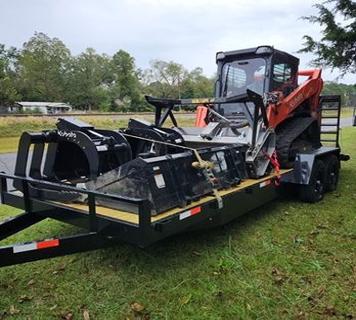 Orange skid steer on a flatbed trailer in a grassy outdoor area.