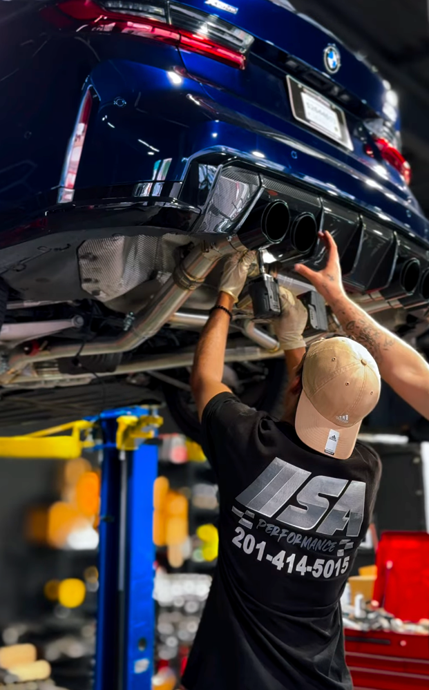 Mechanic working under a blue car on a lift, tightening the exhaust system in a garage | SA Performance