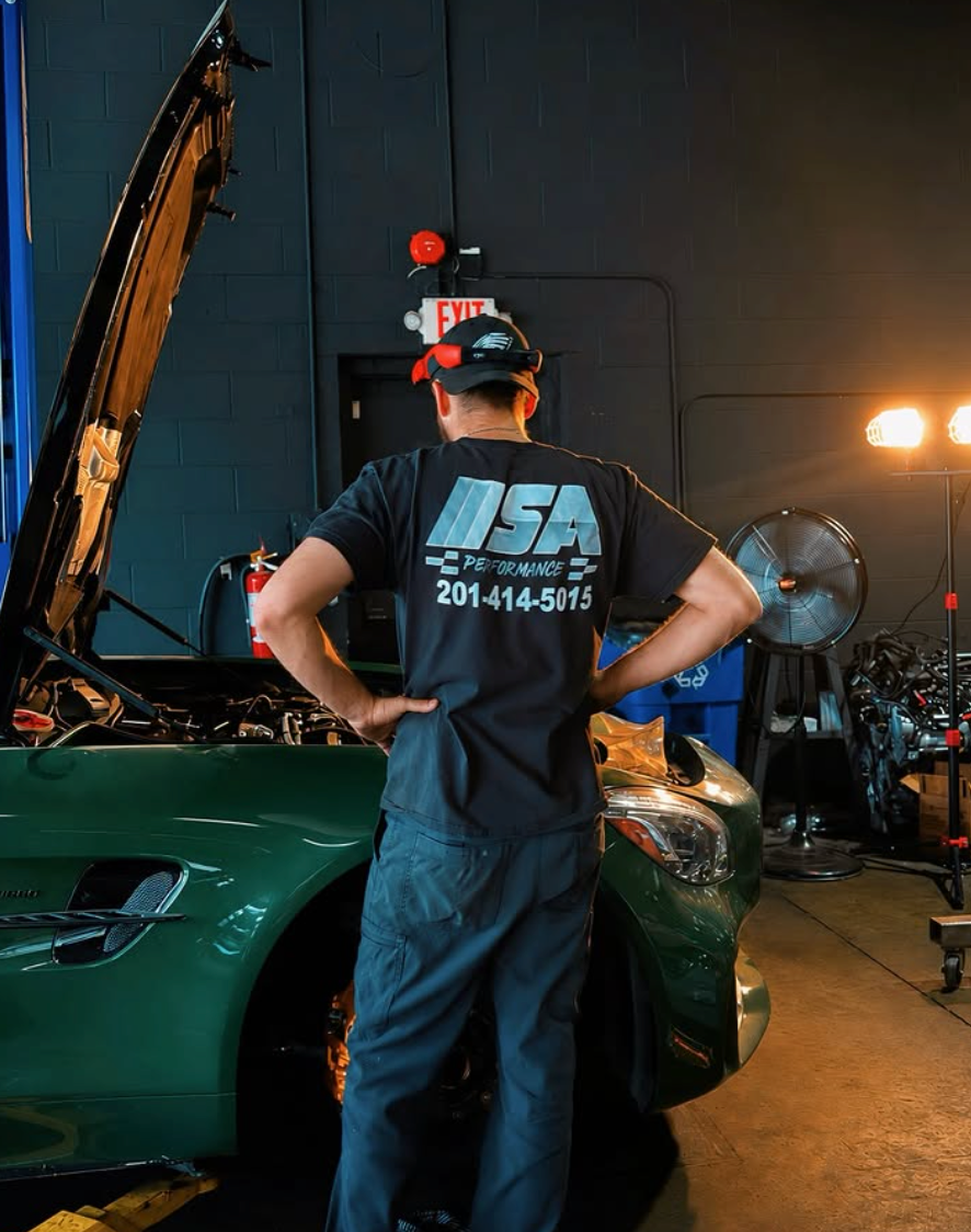 Mechanic standing beside a green car in a dim garage with the hood open | SA Performance