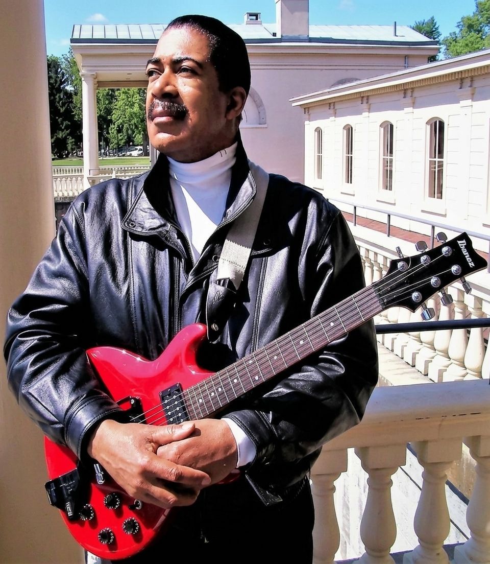 Man standing on a balcony holding a red electric guitar, wearing a black leather jacket, with buildings behind him