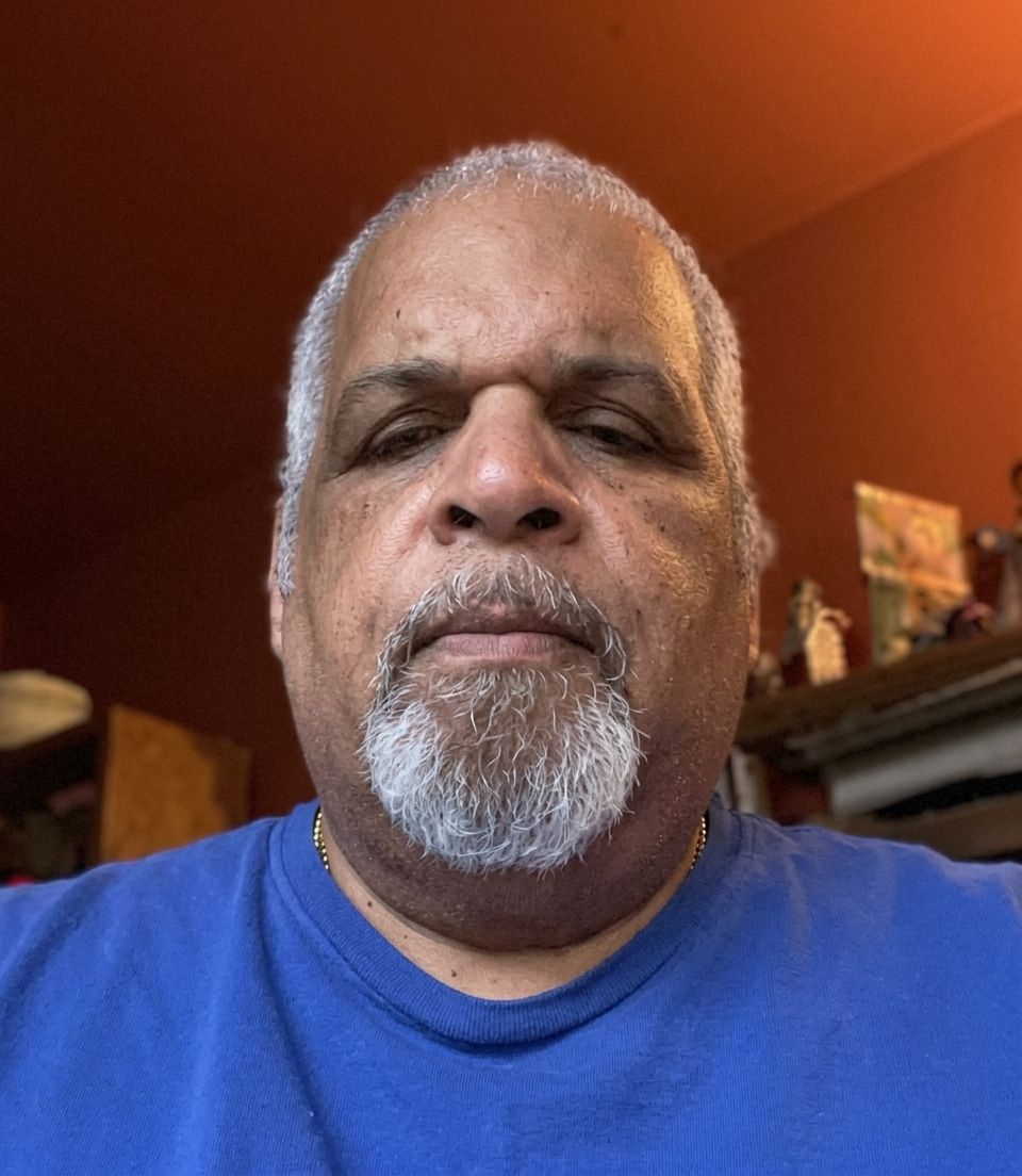 Man in blue shirt and gray goatee looking at the camera indoors against a warm orange wall