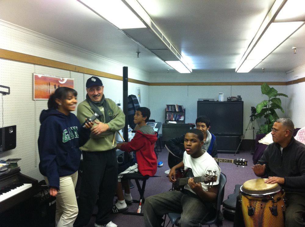 Several people playing music together in a rehearsal room, with a saxophone and conga drum.