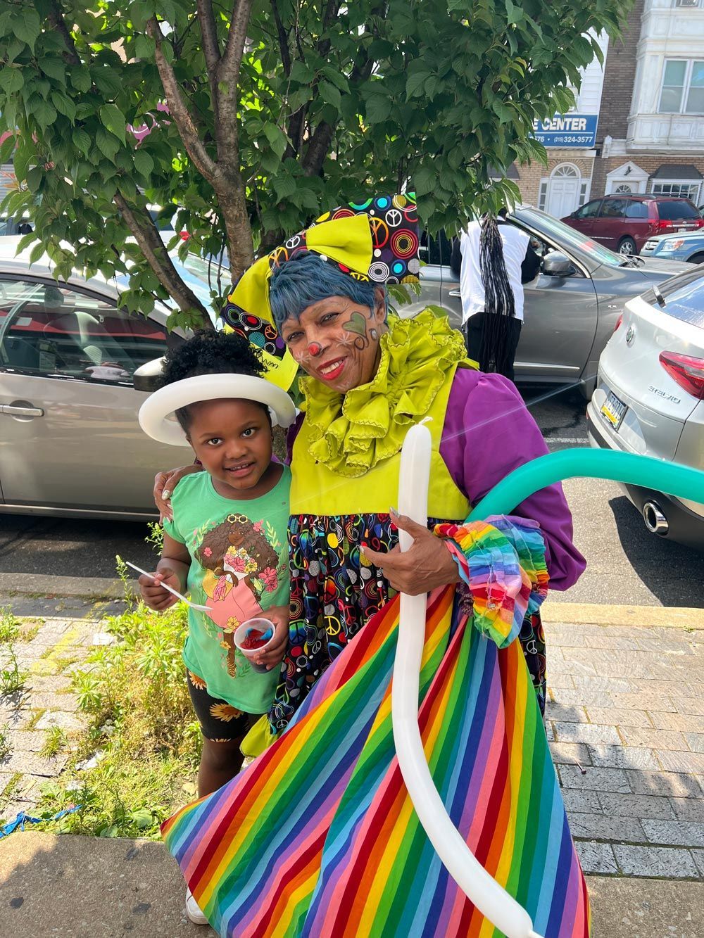 Two people smiling outdoors, holding rainbow-colored fabric, beside parked cars and a tree.