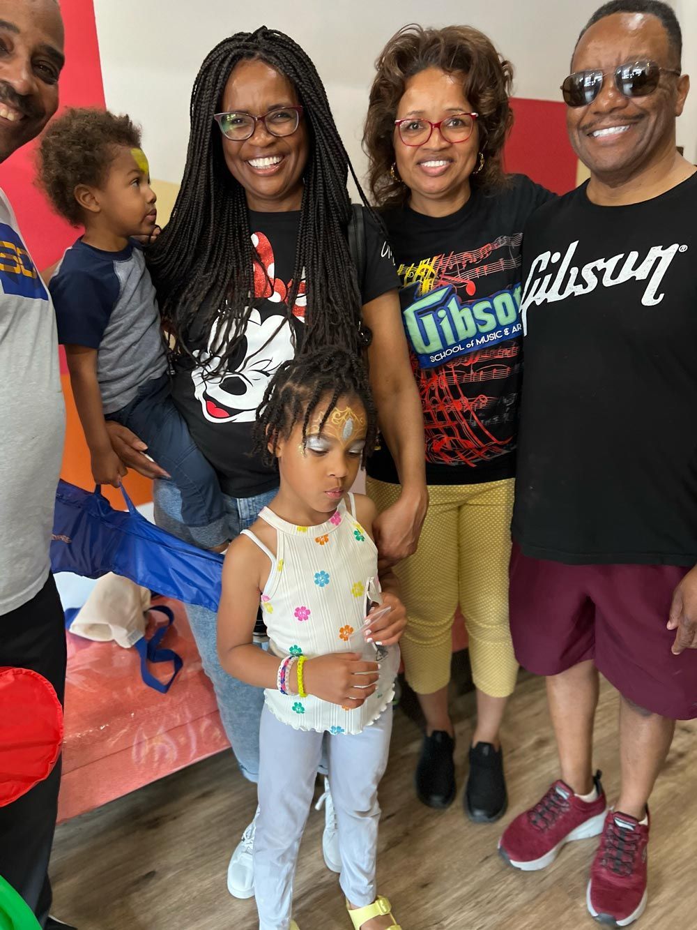 Group of five smiling people posing indoors, with children in front and a black-and-red wall behind them