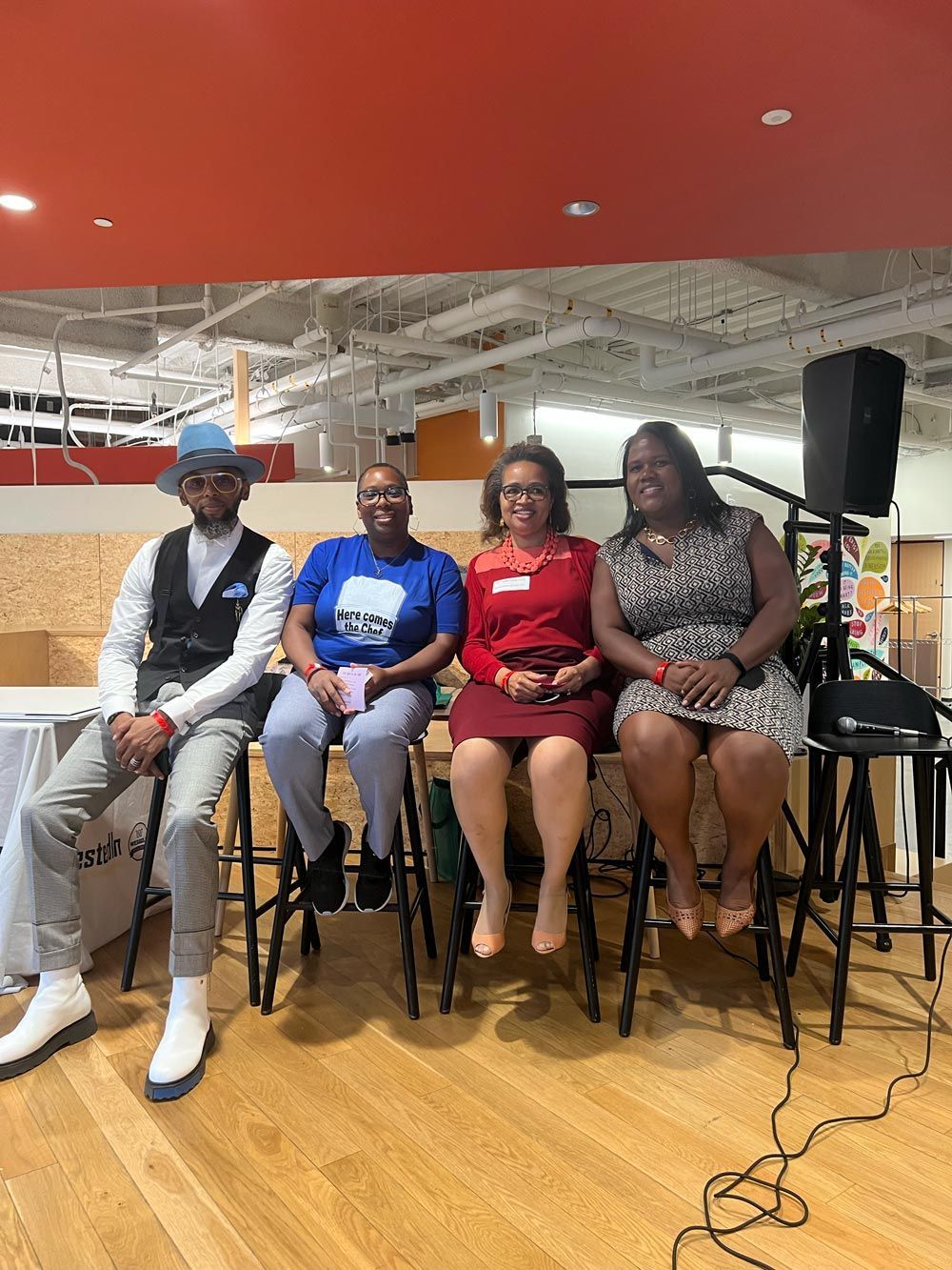 Four people seated on chairs at an indoor event, with a microphone and speaker nearby.