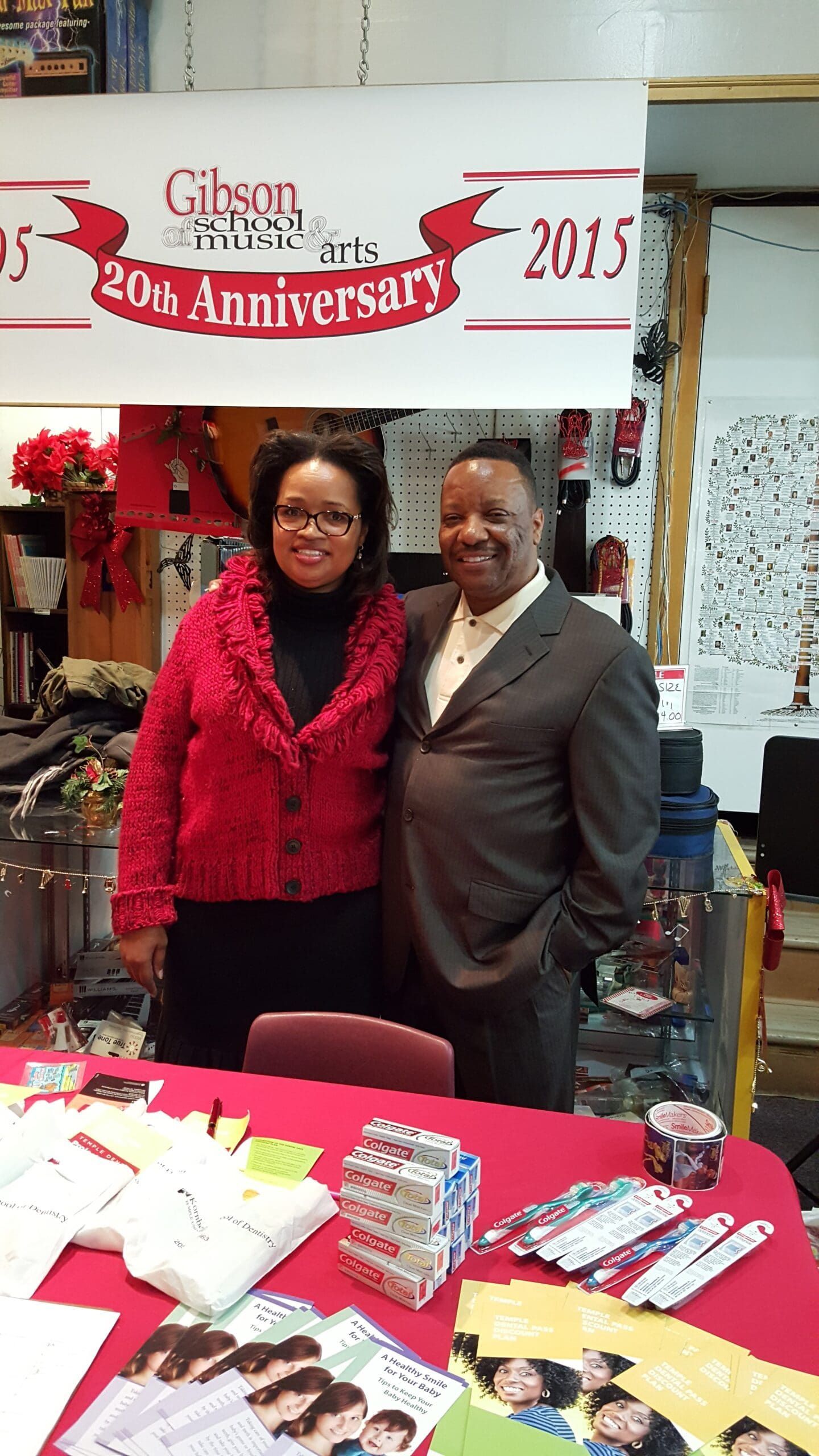 Two people standing behind a table at a 20th anniversary booth with gifts and brochures, smiling.