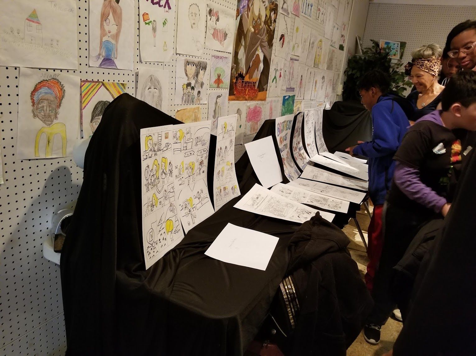 People viewing posters and drawings displayed on a table at an indoor art exhibit