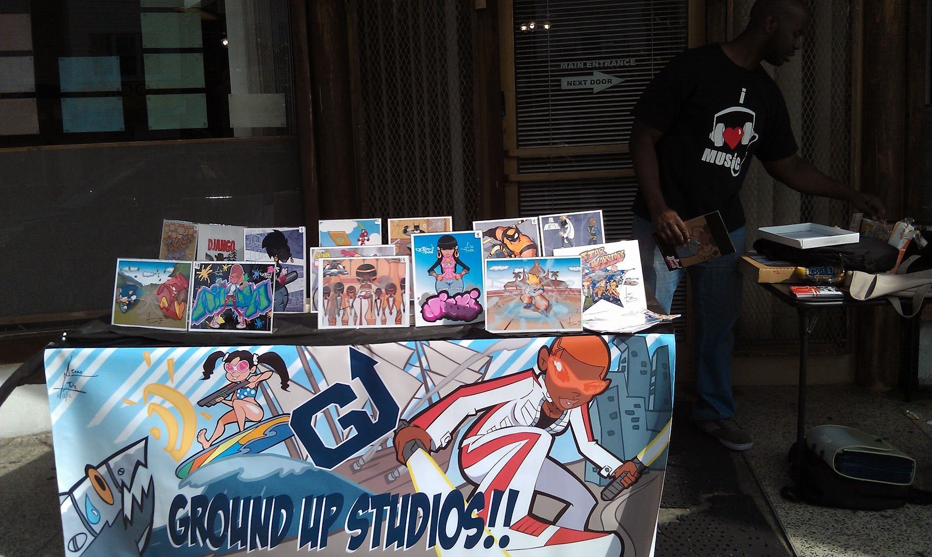 Comic art booth with manga prints and a “GROUND UP STUDIOS!” banner; person stands nearby.