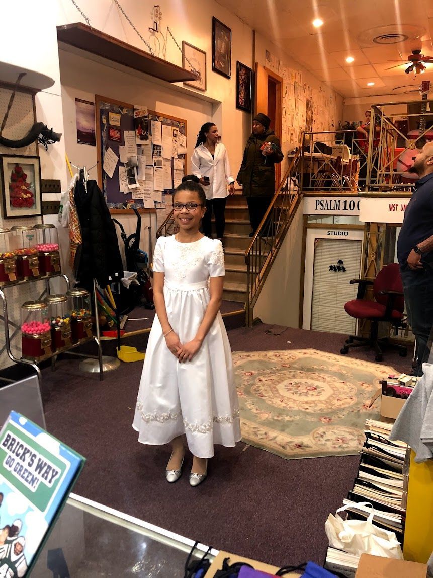 Child in a white dress standing in a vintage-style room with rugs, shelves, and decor.