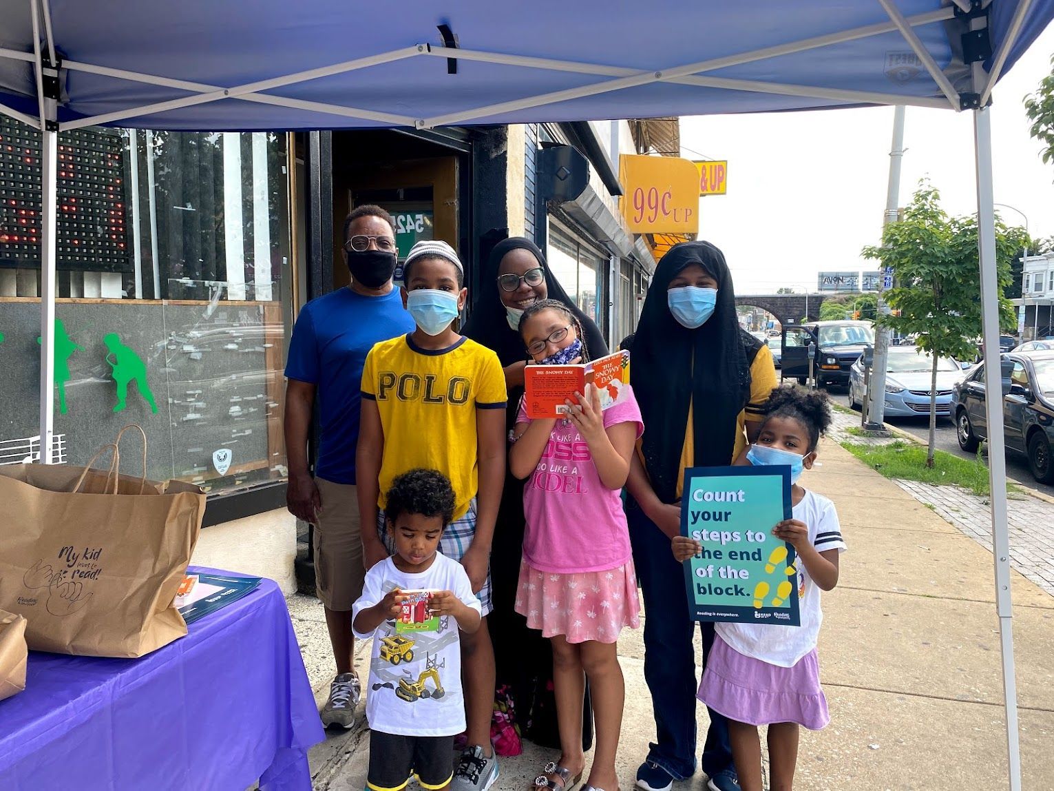 Group holding signs under a blue canopy on a sidewalk, with a purple table and bags nearby.