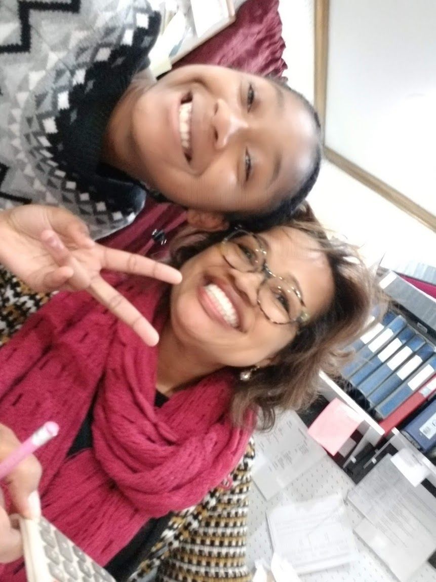 Two smiling people posing indoors, one making a peace sign, with shelves and binders in the background.