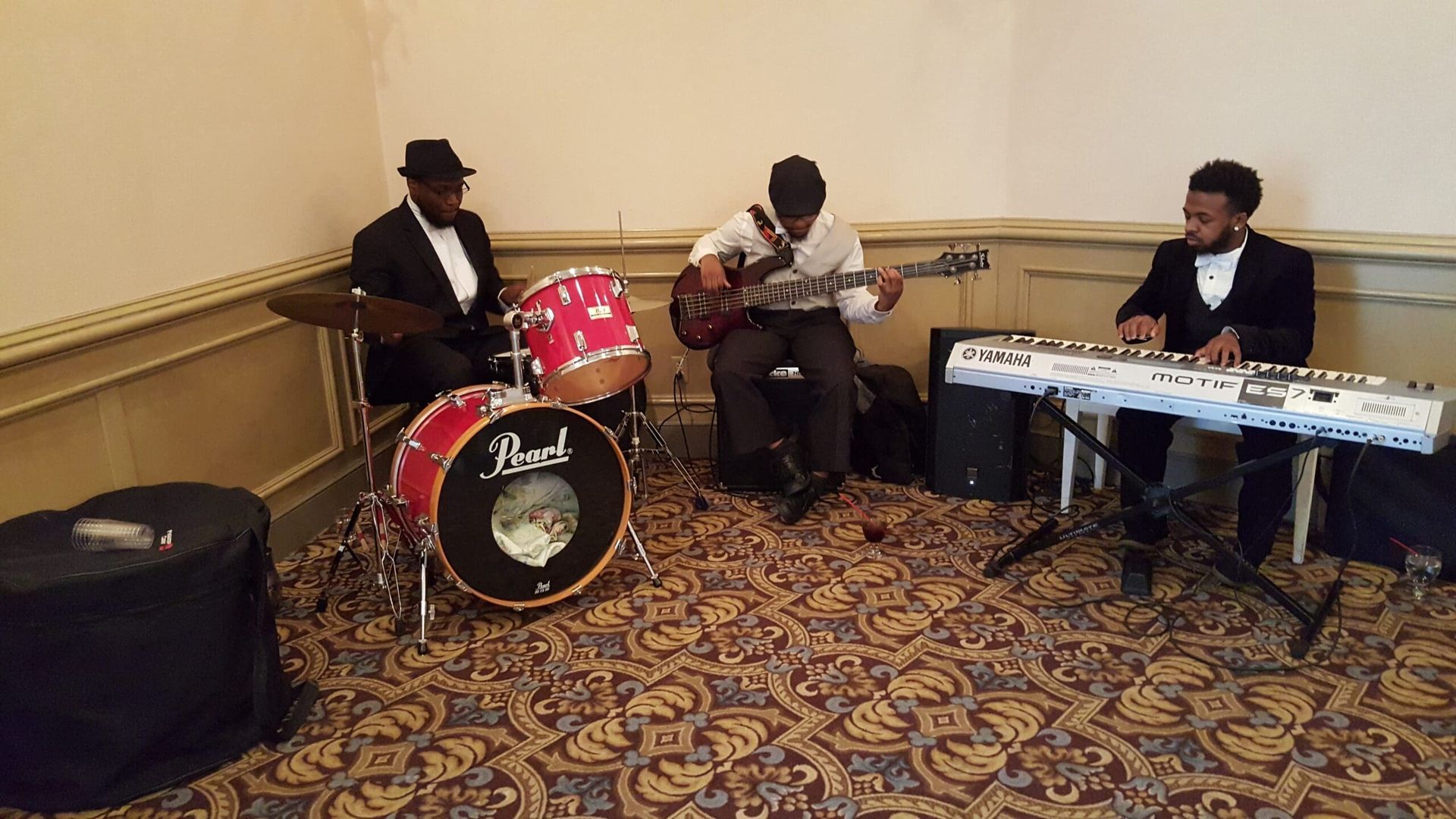 Three musicians perform indoors with drums, guitar, and keyboard in a carpeted room.