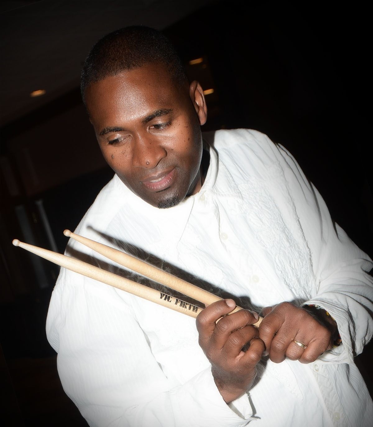 Man holding drumsticks, looking down, in a white shirt against a dark background