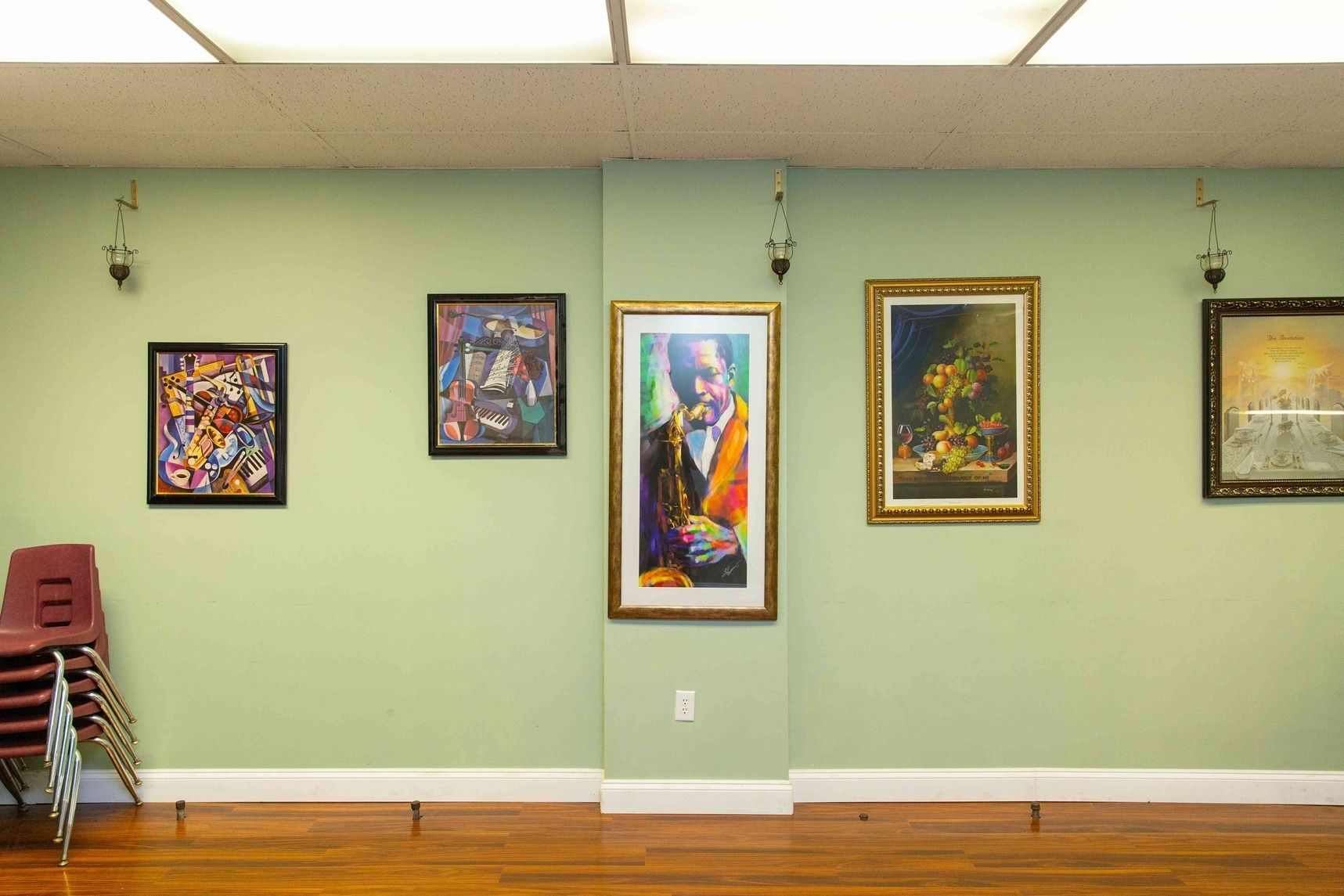 Art gallery wall with five colorful framed paintings on a pale green wall, wooden floor, and stacked chairs at left
