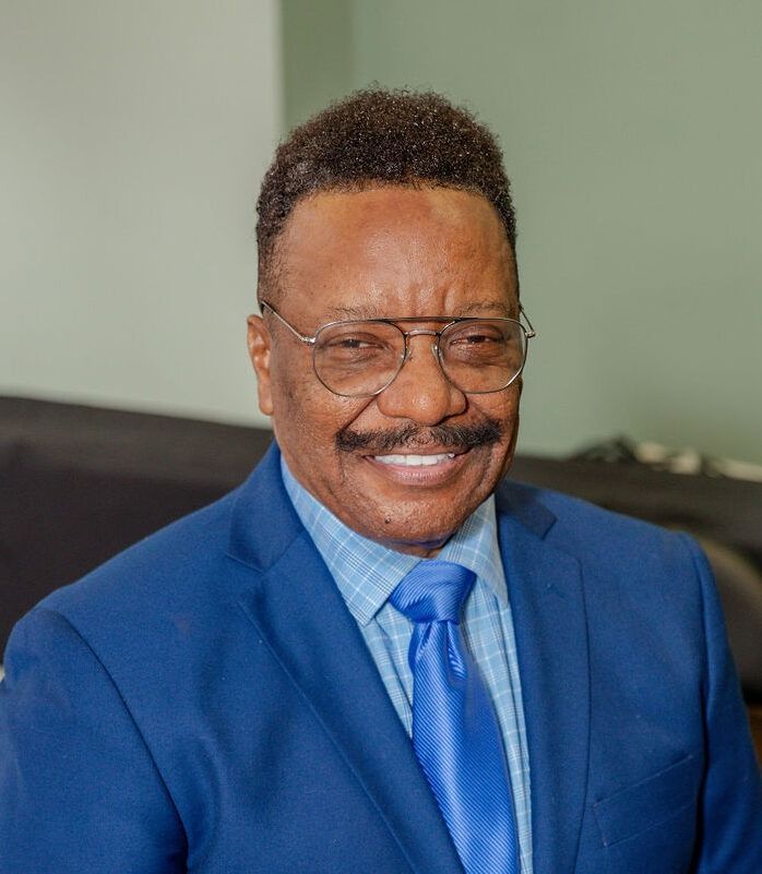 Smiling man in a blue suit, tie, and glasses, seated indoors against a muted green background