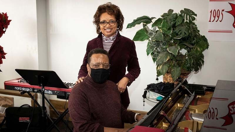 Masked pianist at a keyboard with a woman standing behind, in a room with a plant and music equipment.