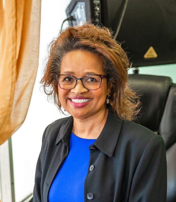 Smiling woman in glasses and black blazer standing indoors beside a window with beige curtains.