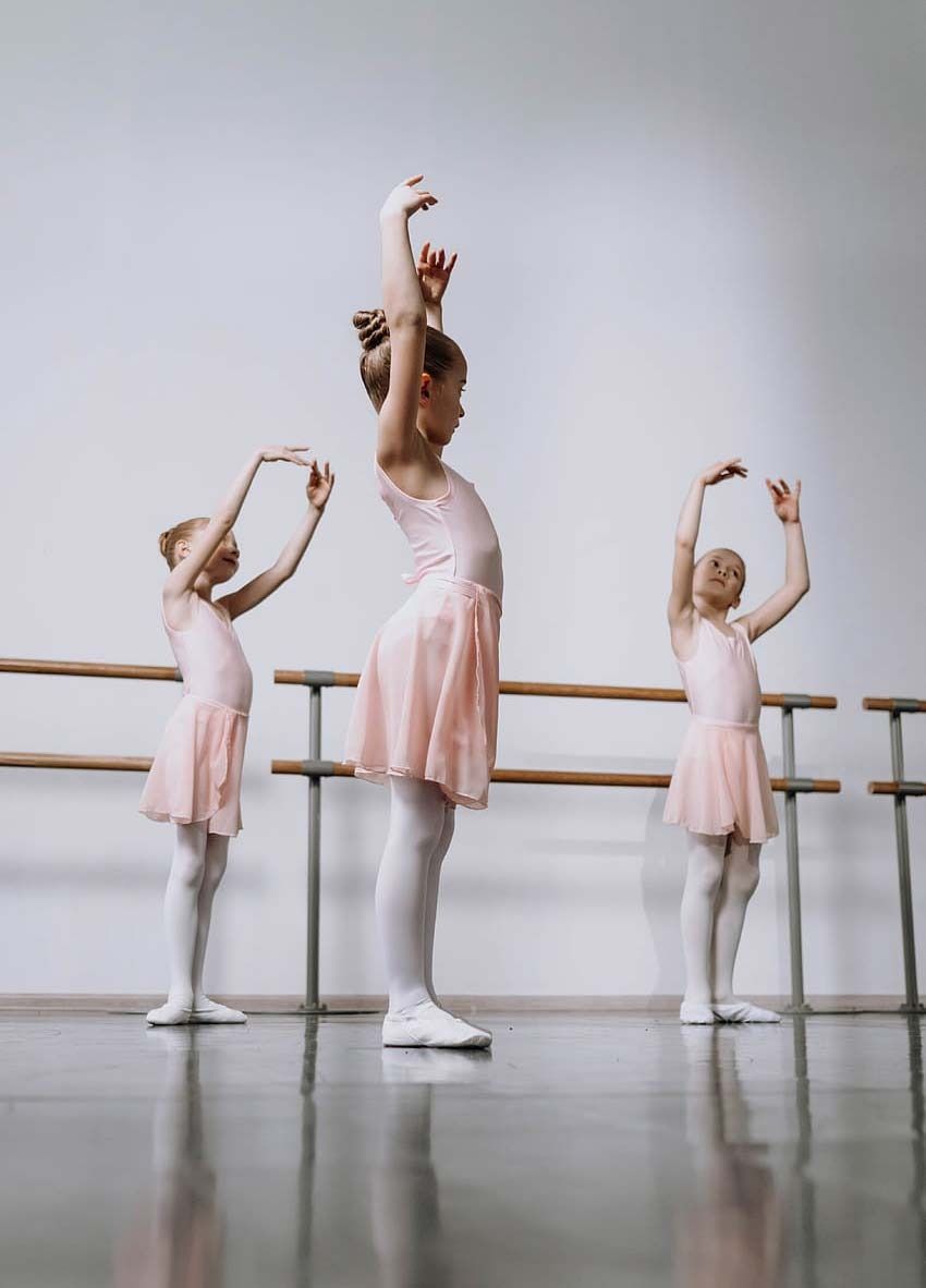 Three ballet dancers in pink leotards and white tights practice arm positions at a barre in a studio.
