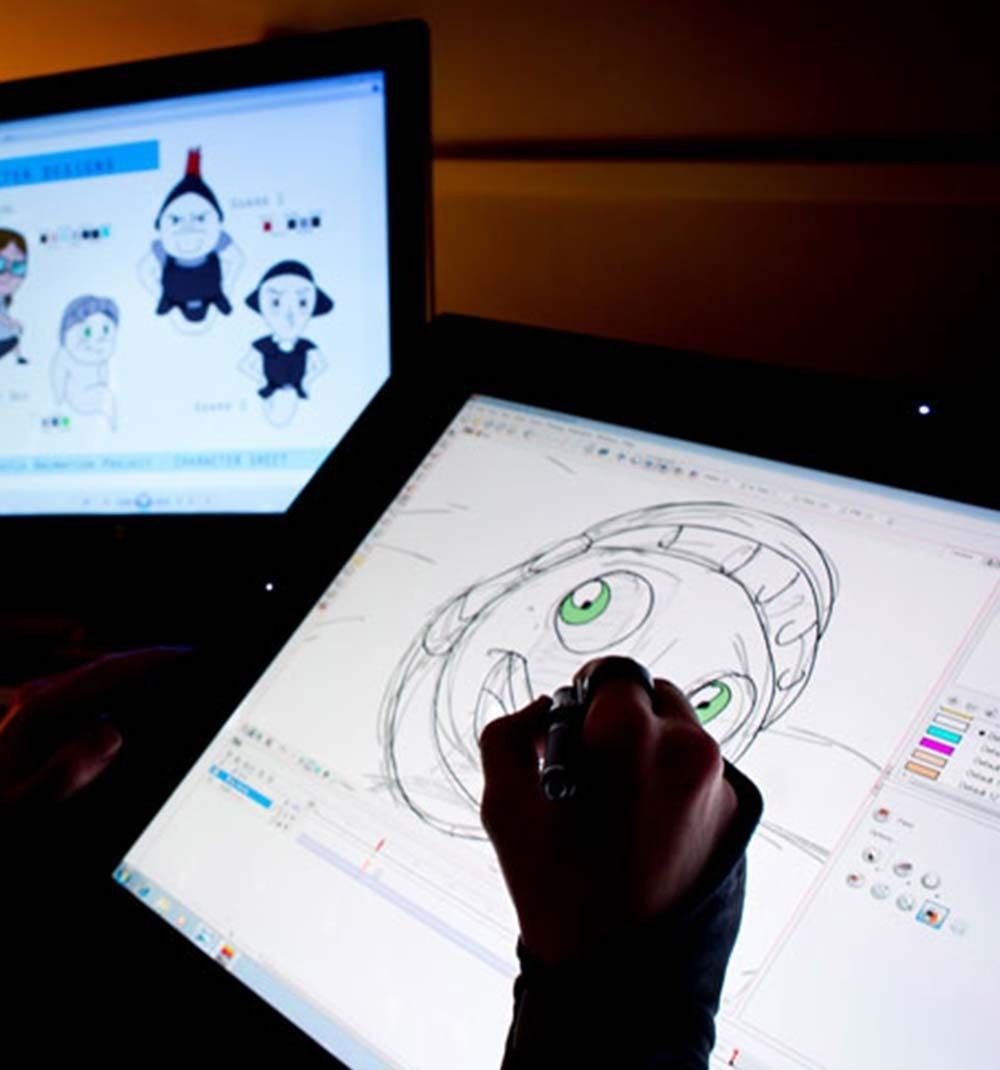 Hand drawing a cartoon face on a digital tablet beside a monitor showing character sketches