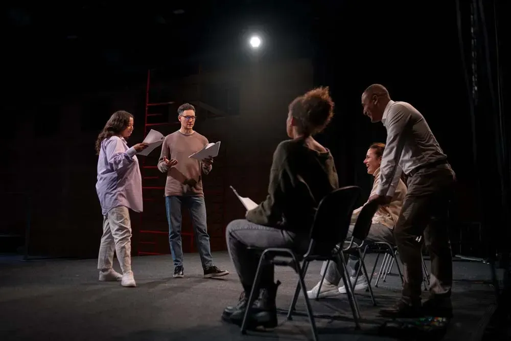 Actors rehearsing on a dim stage under a spotlight, with scripts and chairs arranged in a circle.