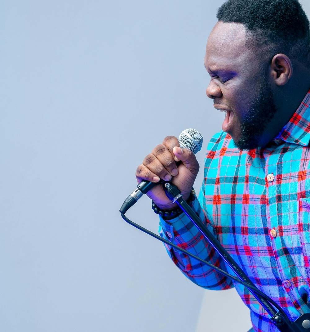 Singer in a blue and red plaid shirt holding a microphone and singing against a plain background