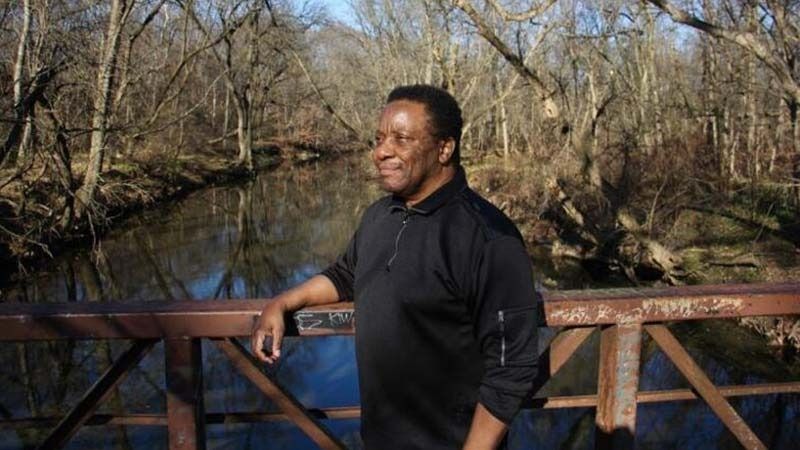 Person leaning on a wooden bridge railing beside a creek in a wooded park