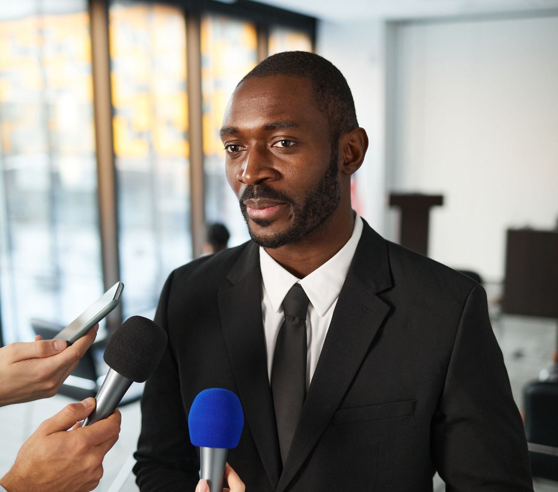 Man in black suit being interviewed by reporters with microphones in an office setting