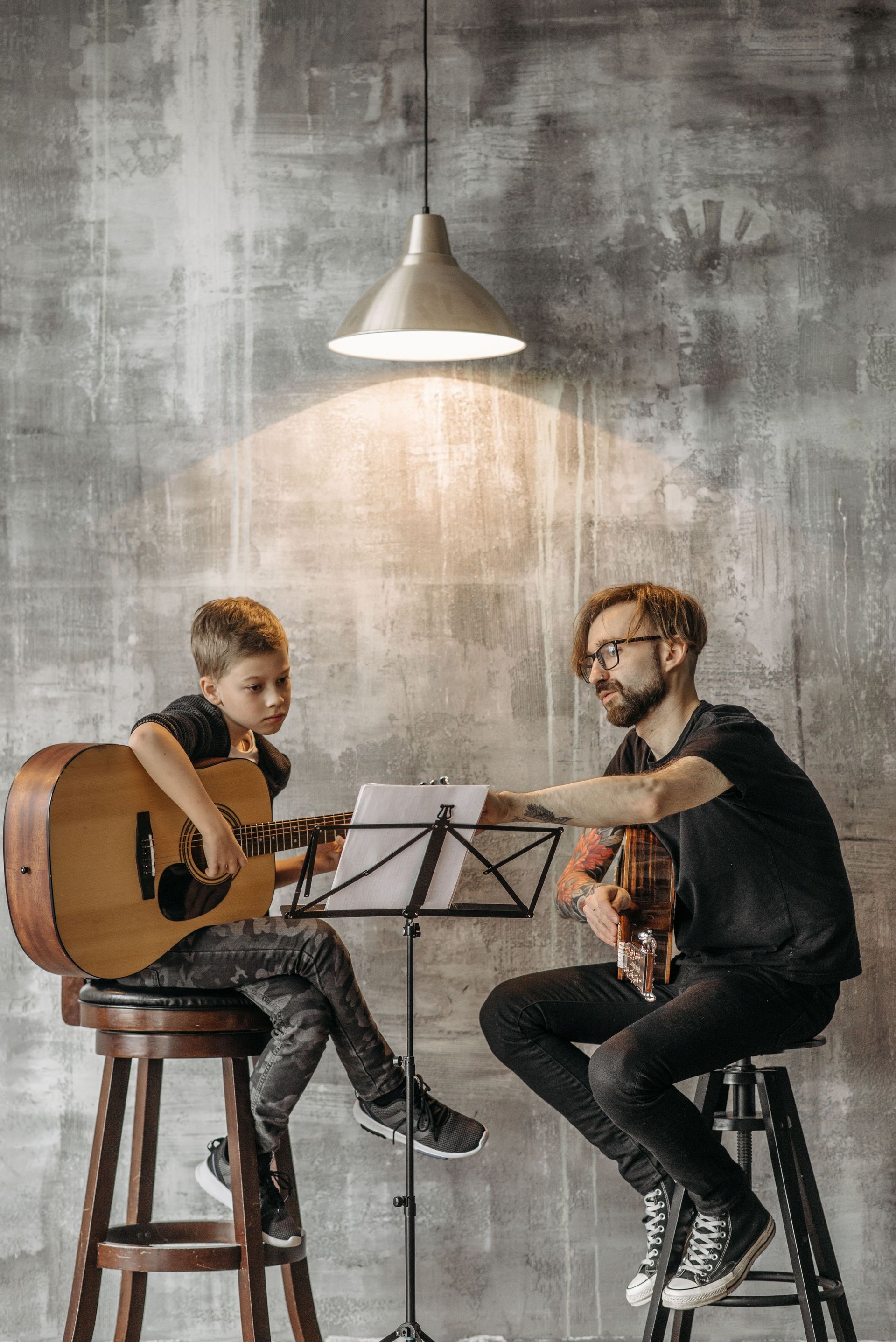 Two musicians seated under a hanging lamp, playing acoustic guitars in a rustic studio.