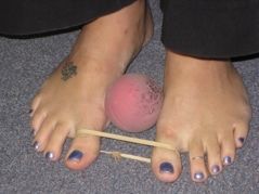 Feet with a pink ball between them, secured with rubber bands, on a gray carpet.