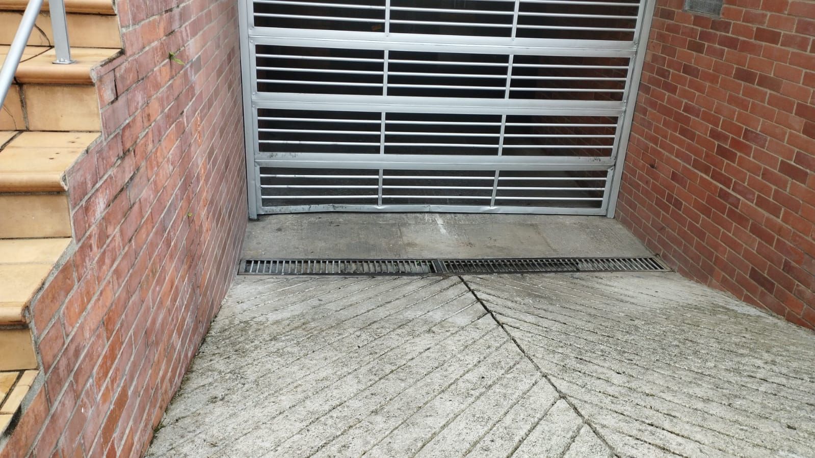 Un camino de hormigón conduce a una puerta de garaje metálica cerrada, flanqueada por muros de ladrillo y una escalera a la izquierda.