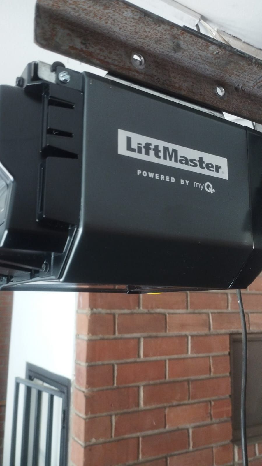 Un abrepuertas de garaje LiftMaster negro montado en una viga de madera del techo, situado frente a una pared de ladrillo rojo.